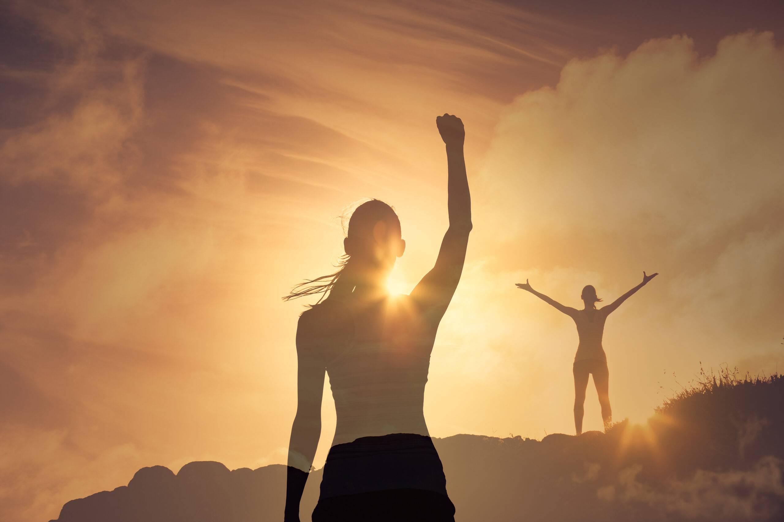 two women scaling a mountain with their hands up in victory and power with a sunset in the background